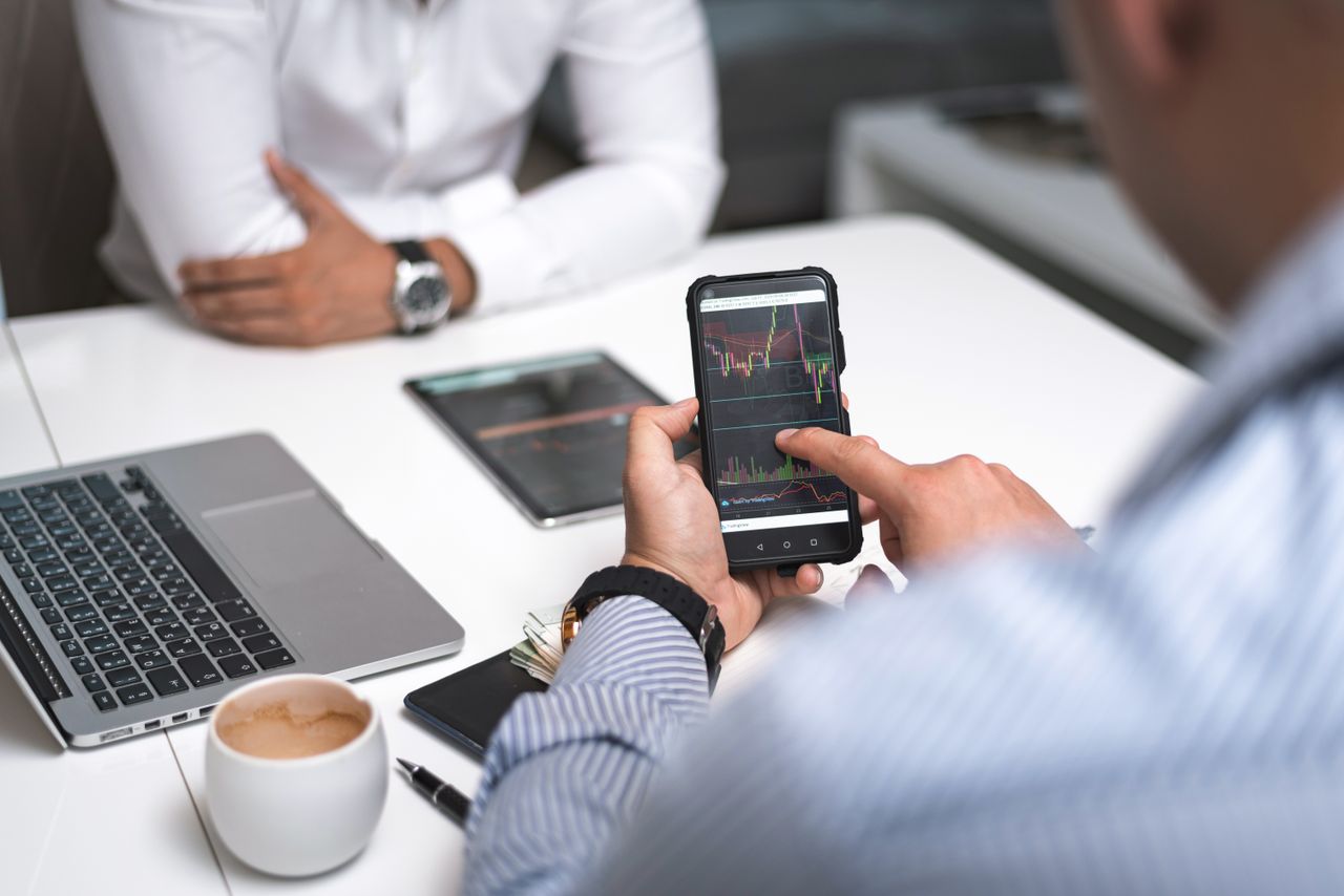 Two businesspeople review stock market figures with a smartphone and laptop in a contemporary office environment.